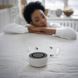 Woman resting on a bed beside the white SIBŌ singing bowl sound bath device
